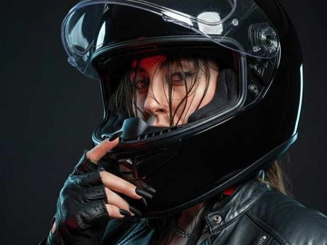 KimyR with a black helmet, wet hair, and black leather jacket, holding the helmet's chin strap, showing black nails, and a serious face.