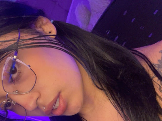 KatianPierce with black hair, glasses, and nose piercing, biting her lower lip, hand in hair, purple lighting, tattoo on right arm, looking seductively at camera.