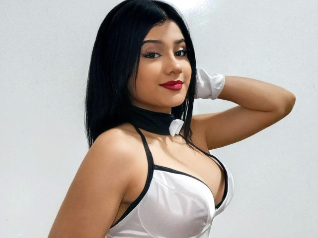 SuesseKlara-hot with black hair, red lipstick, white and black outfit, gloves, and black collar, showing off her tits, looking at camera with a slight smile, arm raised.