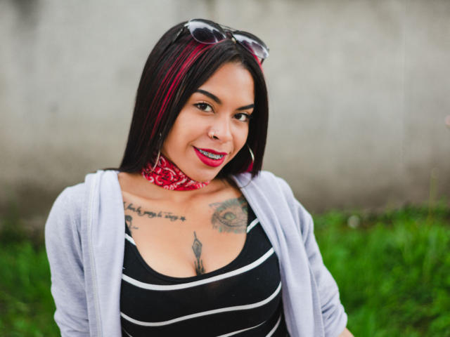 MissRuuby has dark straight hair with red highlights, wearing a red bandana and large hoop earrings. She smiles with red lipstick, showcasing tattoos on her chest and upper arms. She's in a black and white striped top, with a light gray cardigan. Her skin is medium brown, and she has a nose ring. The grassy background is blurred. The image has a natural, candid feel. Her hair and tattoos are prominent. She's centered, with a slight tilt to her head.