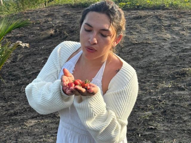 TommyTS with closed eyes, holding red strawberries, wearing a white lace top and chunky white sweater, standing in dirt, green foliage in background.