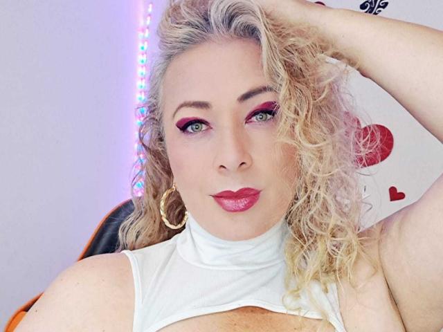 SofiaCurvyCute has curly blonde hair, red eyeliner, and pink lipstick, wearing a white top that exposes her tits. She's looking at the camera with a hand in her hair.