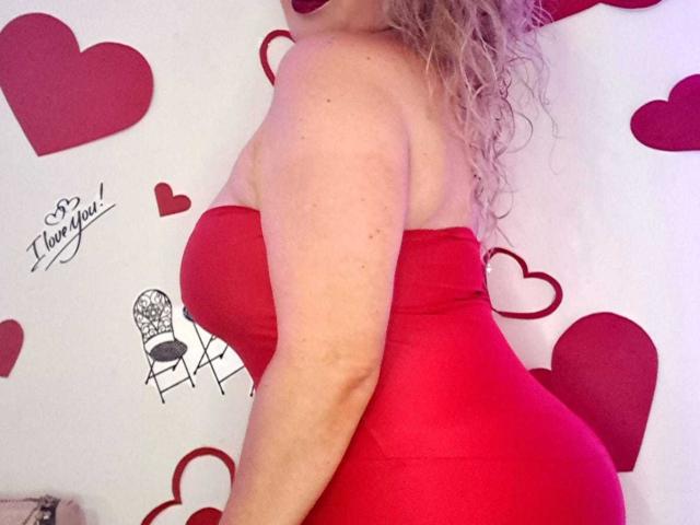 SofiaCurvyCute with curly blonde hair wearing a tight red strapless dress, highlighting her big ass and medium-sized boobs. Red hearts and signature on the background.