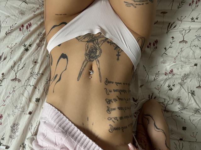 SensualMila's white panties, showing a tattooed flat stomach with a pierced navel, and pink shorts. She has tattoos on her hips and belly, lying on floral bedspread.