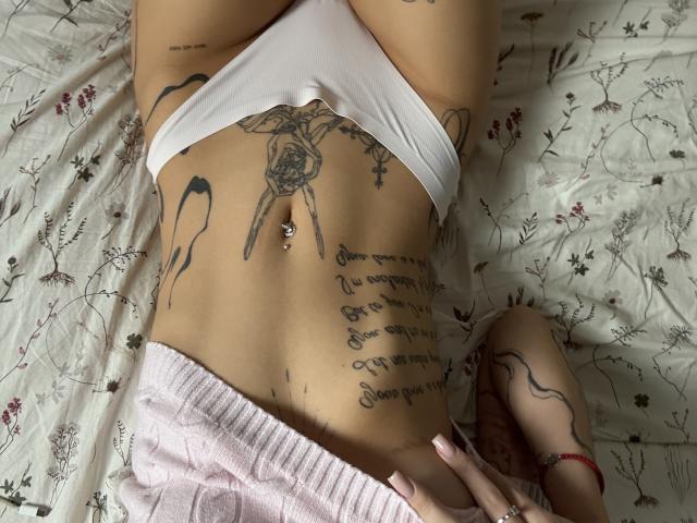 SensualMila's lower torso in white panties, showcasing tattoos, a navel piercing, and a pink ribbed shirt. Her hand rests on her hip, displaying a red bracelet.