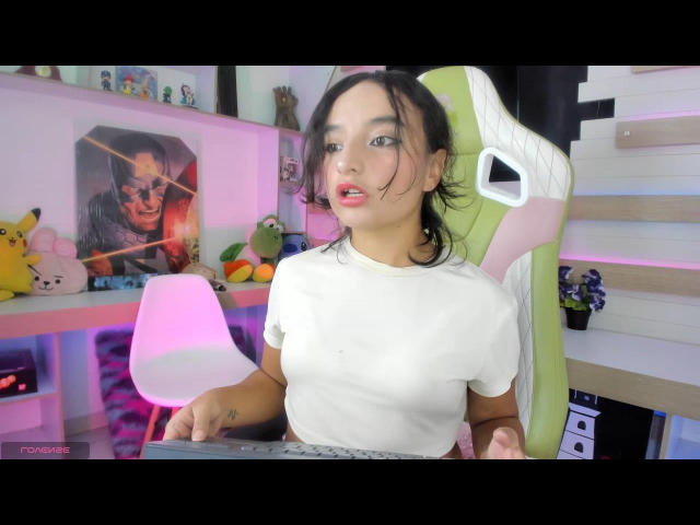 EmilyJonexs with small tits, wearing a white shirt, sitting in a green and white chair, looking at a camera with pink light, black hair, and a slightly open mouth.