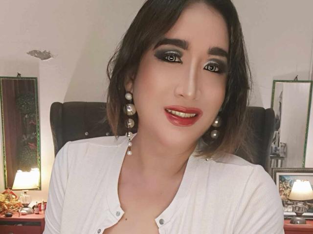 OhhhMyMistress with brown bob, dark eyeliner, red lipstick, pearl earrings, and white buttoned shirt, smiling, showing her tits slightly through shirt.
