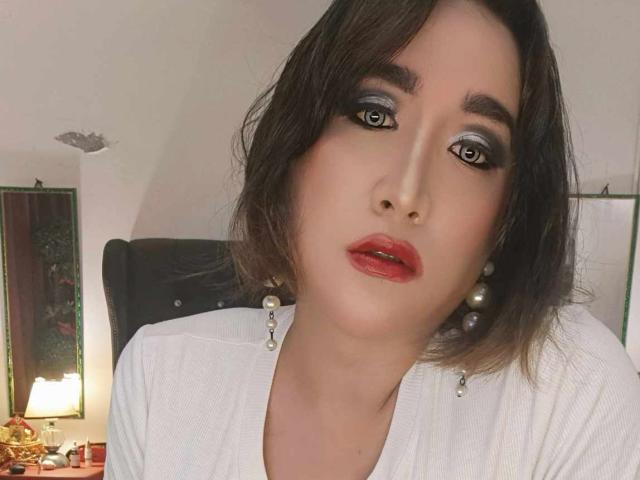 OhhhMyMistress has dark hair, heavy eyeliner, blue eyeshadow, red lipstick, pearl earrings, and a white top, with a slightly parted red mouth.