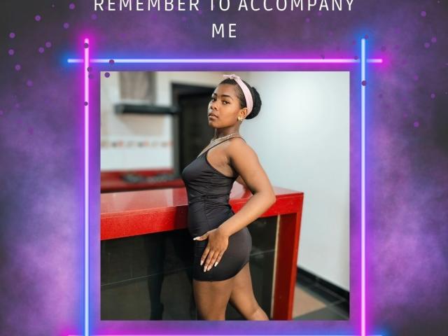 KarinaFlowers with dark skin, wearing a black tank top and shorts, pink headband, standing by a red table, hands on her ass, looking over her shoulder.