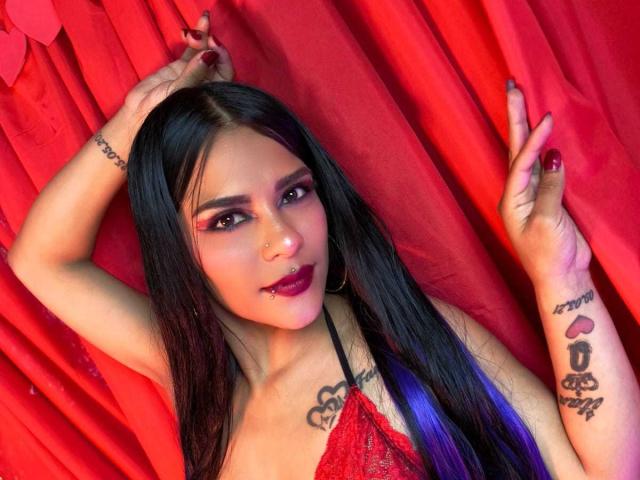 MiaBellucci with long black hair, red makeup, and hoop earrings, wearing a red lace top, showing tattoos on arms, red nails, and a nose piercing, against red curtains.