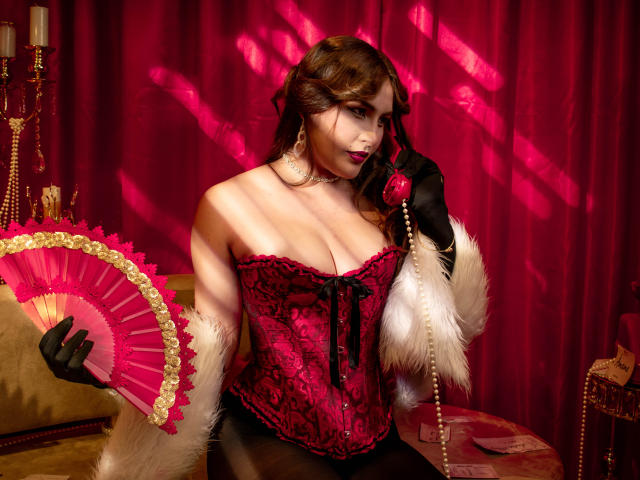 SusyHadid with red lace corset, black gloves, holding pink fan and red phone, white fur cuffs, pink shadowed background, pearls, dark lipstick, wavy brown hair, sexy, busty.