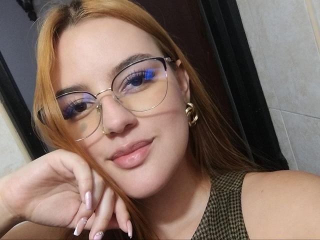 HaileyTims with orange hair, blue eyeshadow, glasses, gold hoop earrings, and pink nail polish, smiling at the camera, resting her chin on her hand.