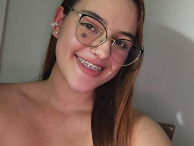 HaileyTims with glasses, nose ring, and earrings, smiling at the camera, showing her teeth with a small black mark under her right eye, topless with long brown hair.