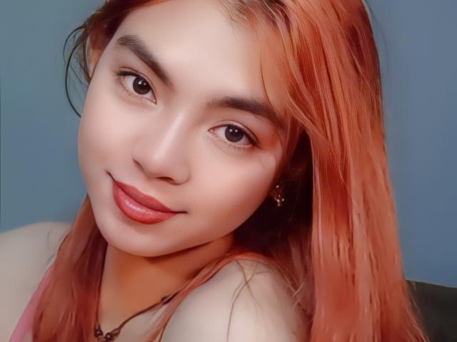 FreakyJane has a cute face with light skin, brown eyes, and orange-dyed hair. She's smiling with pink lipstick and a small nose. She's wearing a necklace.