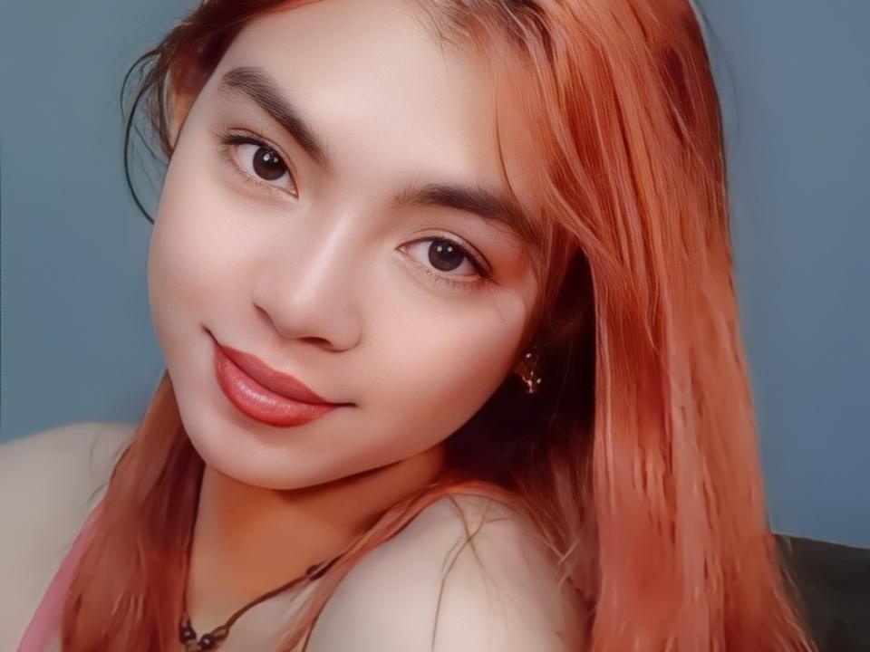 FreakyJane has a cute face with light skin, brown eyes, and orange-dyed hair. She's smiling with pink lipstick and a small nose. She's wearing a necklace.