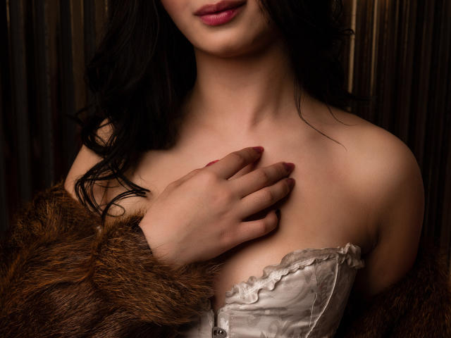 MagieeThomson with dark wavy hair, pale skin, pink lipstick, white strapless top, hand on chest, brown fur coat, visible cleavage, red nails, slightly smiling.