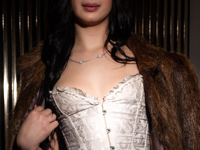 MagieeThompson with long black hair, wearing a white lace corset and brown fur coat, holding it open to show her tits. She has a delicate necklace with a small gem.
