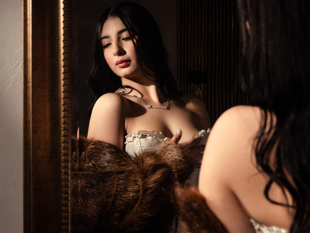 MagieeThompson with dark hair, eyes closed, in a fur coat, exposing her tits, reflected in a mirror, wearing a lace top, necklace, and soft lighting.