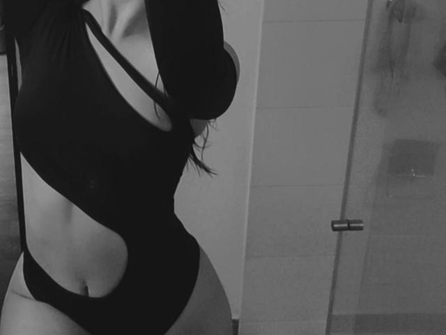 MagieeThompson in a black, cut-out one-piece, showing her slim, curvy body, with visible tits and a small, round ass, standing in a bathroom, photographed in black-and-white.