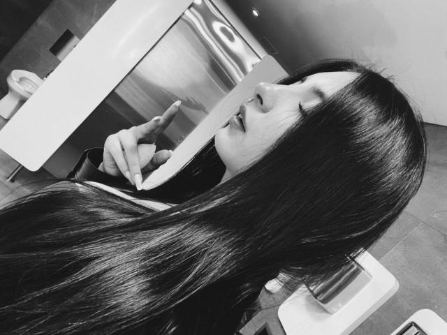 Black-and-white photo of MagieeThomson with long, shiny dark hair, eyes closed, pointing at mirror with right index finger, wearing a dark shirt, in a modern bathroom.
