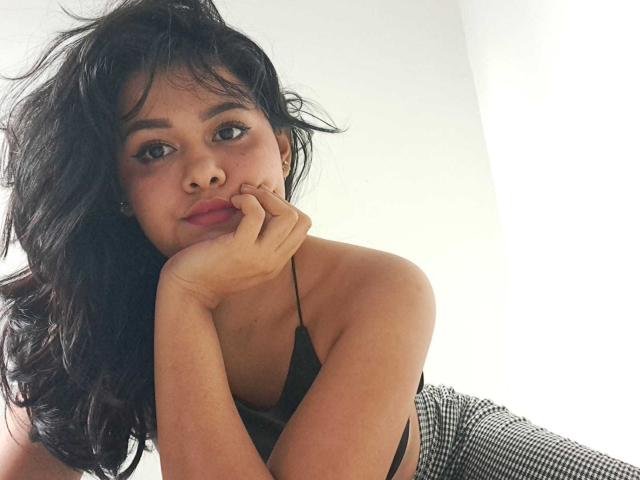 JennLunaLee with messy black hair, resting her chin on her hand, wearing a black halter top and checkered pants, looking at the camera with a subtle smile.