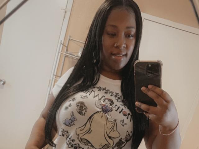 Ebhony with dark skin and long black hair, holding a phone, wearing a white T-shirt showing a graphic of a woman, smiling at her reflection, showing her big tits.