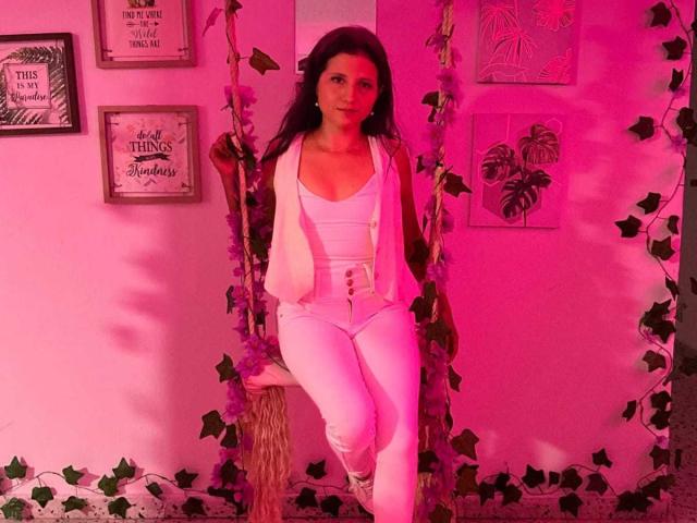 RenataRivas with dark hair, white top, and pants, buttoned vest, sitting on a wooden swing with vines, in pink-lit room, looking at camera, legs crossed.