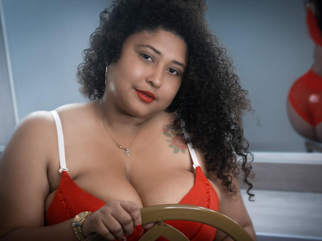 Ayliin, a plus-sized woman with curly black hair, wears a red bra with white straps. She has a tattoo on her left shoulder and is holding a golden chair. Her red lipstick contrasts with her fair skin. She wears a gold bracelet on her right wrist and a small necklace. Her expression is calm and slightly smiling. The image focuses on her upper body. She has large breasts. The background is blurred, with a partial view of a red, curvy object. The lighting is soft, highlighting her facial features and hair texture. The composition emphasizes her upper body and facial expression.