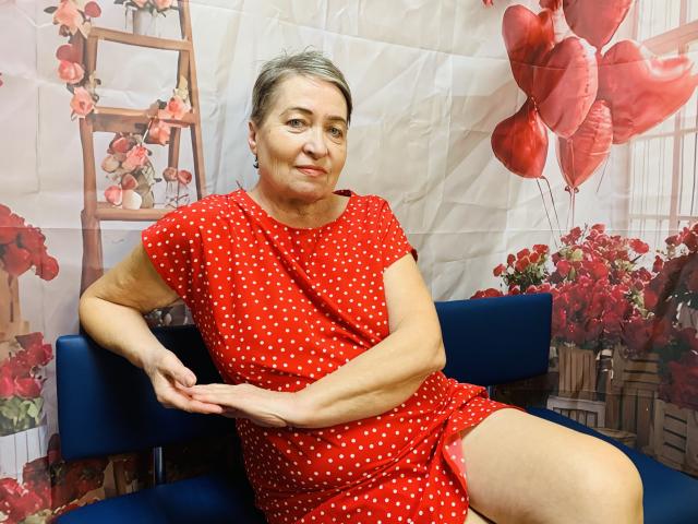 OlgaLuck, una mujer de mediana edad con cabello corto y gris, vestida con un vestido rojo de lunares, sentada en un sofá azul, con las piernas ligeramente separadas, los brazos apoyados en las rodillas, y globos rojos en el fondo..