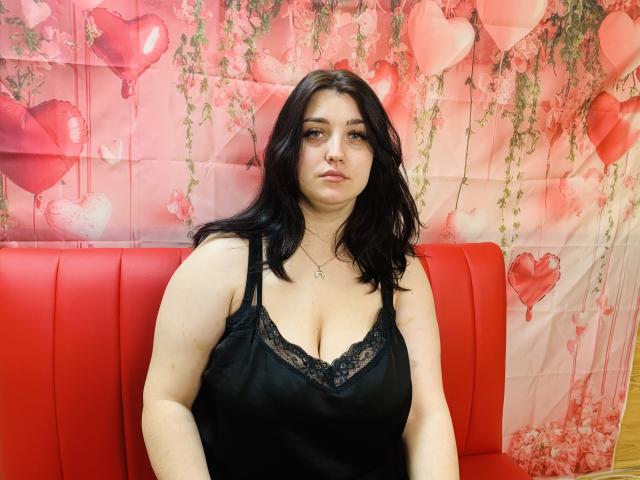 ScarletCargo with black lace-trimmed top, showing her big boobs, sitting on red seat, looking serious, medium-length black hair, fair skin, small nose ring, necklace, soft pink background.