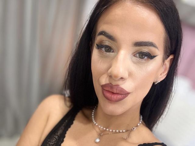 LuxuryAnne has dark hair, heavy eyeliner, and plump maroon lips. She's wearing a black lace top and a pearl necklace. Her skin is light with a subtle sheen.