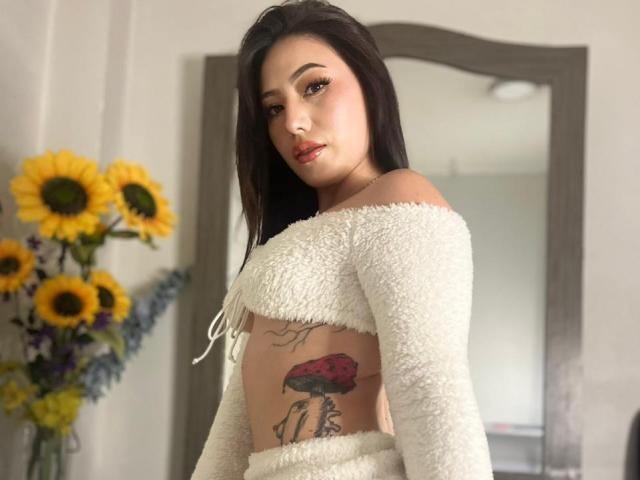MiaParys with dark hair, white fluffy off-shoulder top, red lipstick, tattoo of red flower on her side, standing, looking at camera, sunflowers in background.