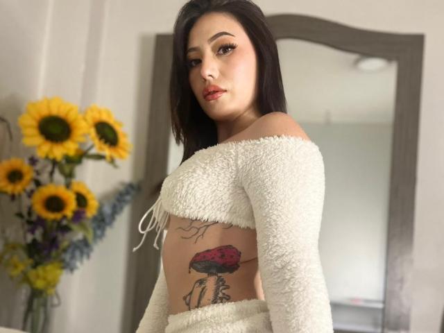 MiaParys with dark hair, white fluffy off-shoulder top, showing a red bird tattoo on her side, standing in front of a mirror, with sunflowers in the background.
