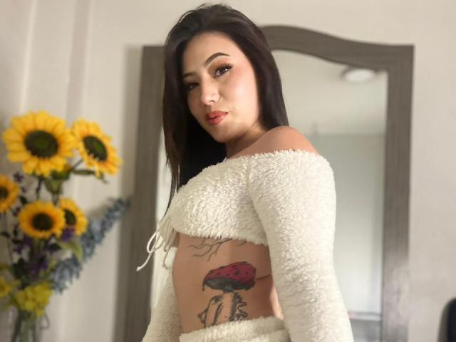 MiaParys with long dark hair, white off-shoulder fluffy top, showing tattoo of red flower on right side, standing in front of mirror, sunflowers in background.