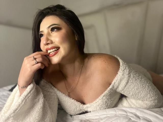 MiaParys, laying on a bed, smiling with her finger on her lip, wearing a fluffy white off-shoulder top, showing her tits, dark hair, and freckles, looking cute.