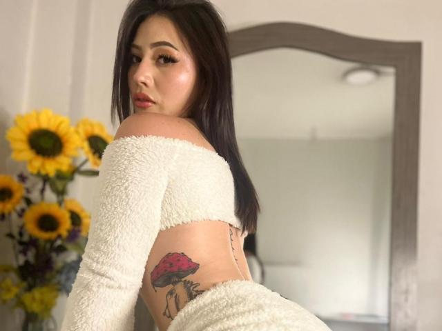 MiaParys has long black hair and is looking over her shoulder with a serious expression. She wears a white, fluffy off-shoulder crop top that exposes her mid-back. She has a colorful tattoo of a red mushroom on her right hip. The background includes a large mirror and a vase with yellow sunflowers. Her makeup is subtle, with pink lips and subtle eyeshadow. The image has a soft, natural light. She has light skin and a curvy, attractive physique.