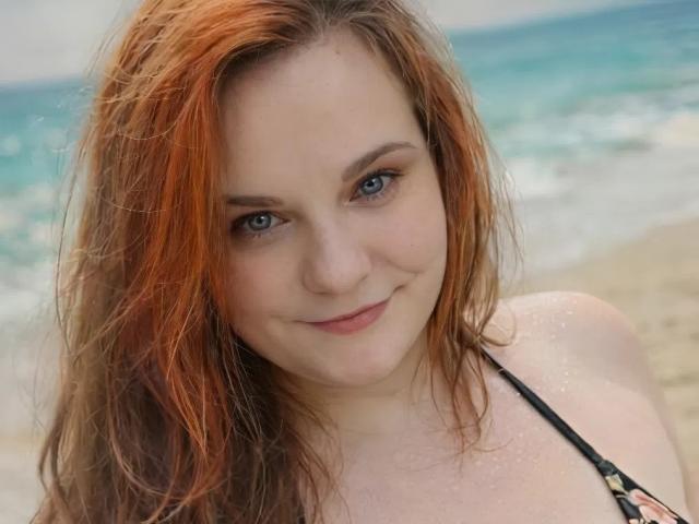 Mavka69 with wet, red-orange hair, light skin, and blue eyes, smiling at the camera, wearing a black halter top, standing on a sandy beach with ocean in background.