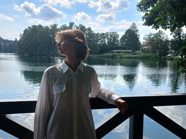 AnnaWestt stands by a black railing, wearing a white, semi-sheer shirt with pockets, her arm resting on it, looking sideways, with a calm, reflective lake and trees in the background.