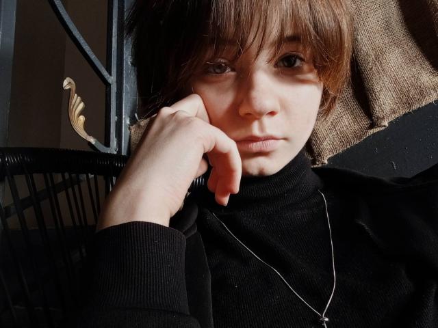 AnnaWestt with brown hair, fair skin, and a serious expression, wearing a black turtleneck and silver necklace, resting her chin on her hand in a dark room.