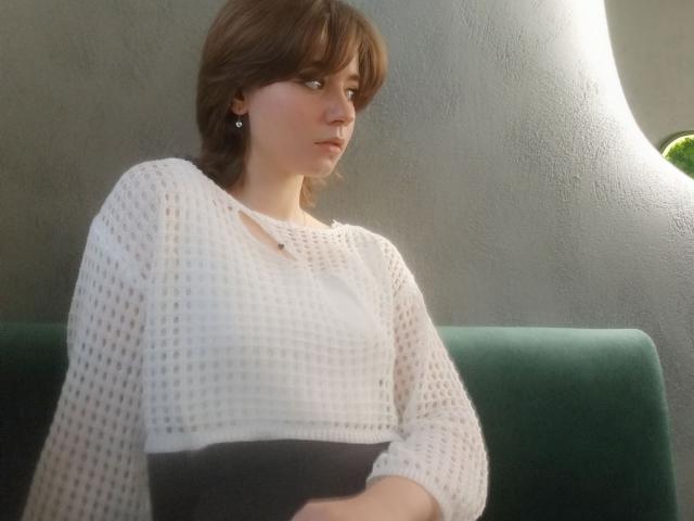 AnnaWestt with short brown hair, white knit top, and small earrings, sitting on green couch, looking to the side, light from the right, hand on black skirt, soft light.