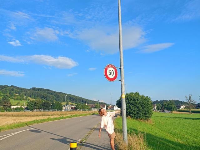 AnnaWestt with short brown hair, wearing a pink shirt and white shorts, leans on a 50 km/h speed limit sign, holding a yellow bucket, standing on a rural road.