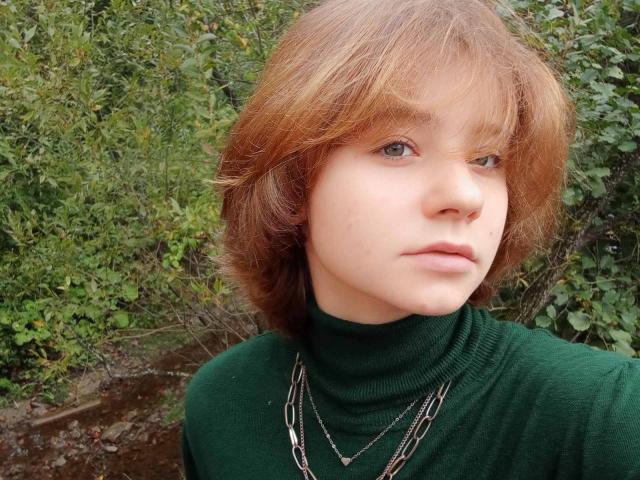 AnnaWestt with red, shoulder-length hair, green turtleneck, and two silver necklaces, looking serious, pale skin, small nose, green eyes, and a slightly pouty mouth.