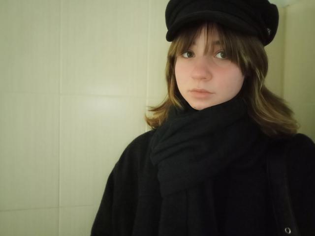 AnnaWestt with pale skin, brown wavy hair, wearing a black hat and scarf, looking serious, black coat, soft light, slightly bluish-gray eyes, subtle pink lips, neutral expression.