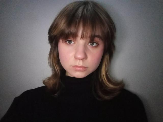 AnnaWestt has light brown hair with bangs, green eyes, and pink cheeks, wearing a black turtleneck, staring straight at the camera with a neutral expression.