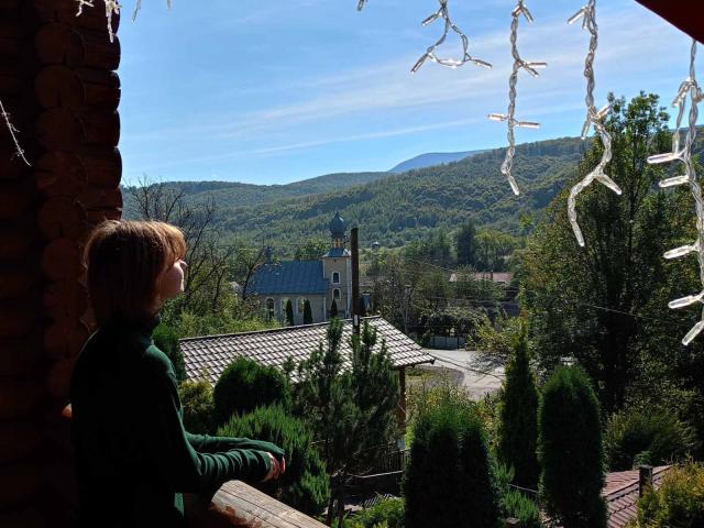 AnnaWestt with short brown hair, wearing a green sweater, standing on a wooden balcony, gazing at a distant church and hill under a bright blue sky.