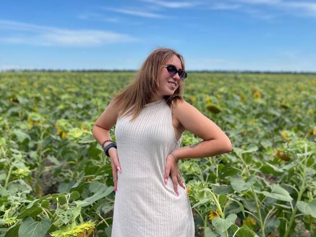 Kamella, wearing a white sleeveless dress, stands in a sunflower field with hands on hips, brown hair, sunglasses, and a smile, showing her ass in the photo.