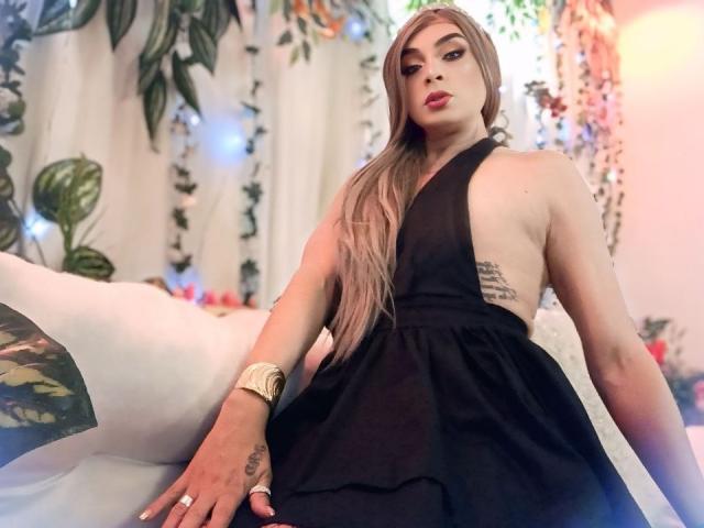 KarolElegance sits on a bed, wearing a black halter dress that reveals a tattoo on her right side. She has long, straight, blonde hair, bold red lipstick, and a gold watch on her left wrist. Her right hand, with white-painted nails, rests on the bed. The image focuses on her confident, slightly tilted pose. Her skin is fair, and she has a subtle, attractive, feminine look. The background includes leafy plants and soft, warm lighting.