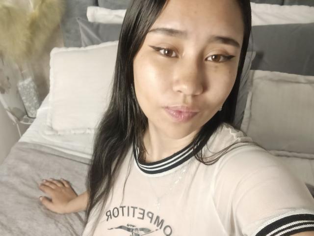 Karollteyes with long black hair, wearing a white shirt with black stripes, pouting her glossy lips, sitting on a bed with white pillows and gray blanket.