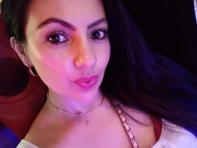 Close-up of JakyKidman with medium skin tone, dark straight hair, and prominent eyebrows. She has pink lipstick and a silver chain necklace with a small pendant. Her large brown eyes look directly at the camera. She wears a white top with a red strap. The lighting has a pinkish-purple hue, highlighting her facial features. The background is dark and mostly out of focus. JakyKidman's expression is neutral but slightly curious. The image is a selfie.