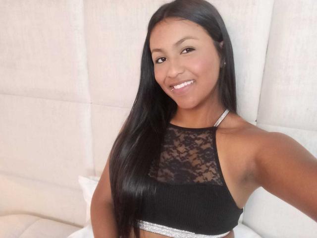 NaomiRios with dark skin and long black hair, smiling, wearing a black lace crop top, showing her small tits and armpits, sitting on a white bed.